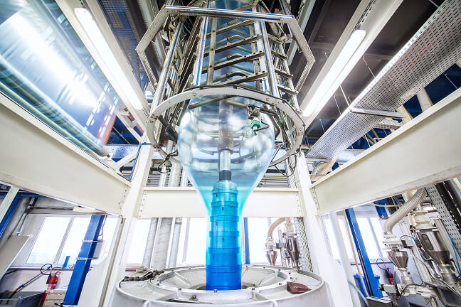 Industrial machinery forms transparent plastic film through a blue extrusion head inside a brightly lit factory hall.