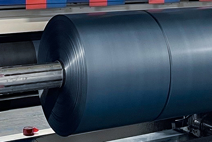Large roll of dark gray plastic film mounted on a metal shaft inside an industrial production machine for processing.