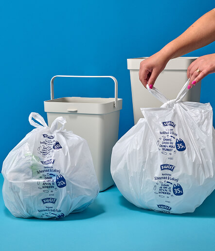 Two printed white trash bags stand beside waste bins while a pair of hands ties the bag on the right closed