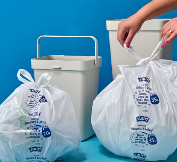 Two printed white trash bags stand beside waste bins while a pair of hands ties the bag on the right closed