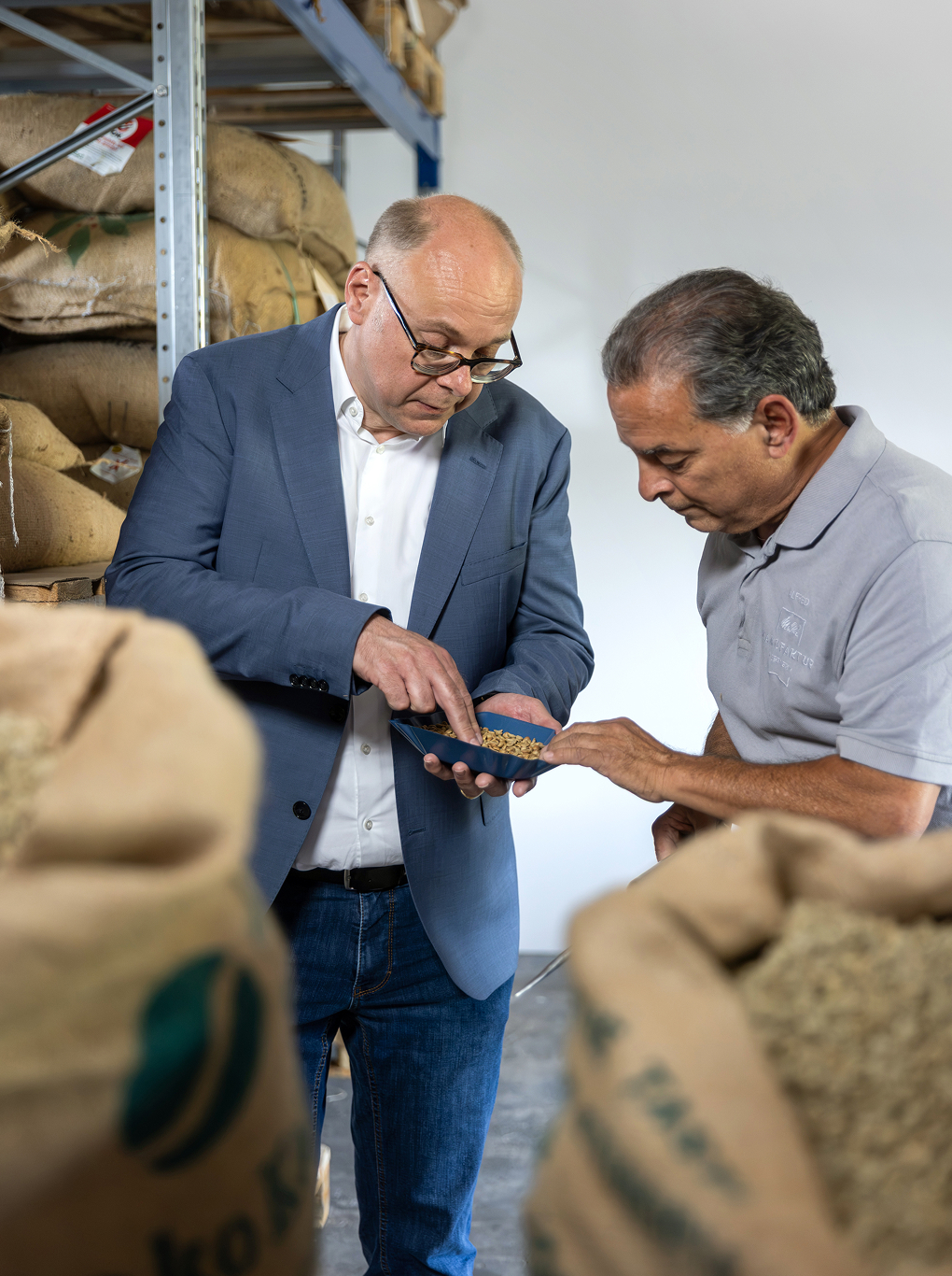 Two men inspect unroasted coffee beans together in a tray among stacked burlap sacks inside a coffee storage warehouse.