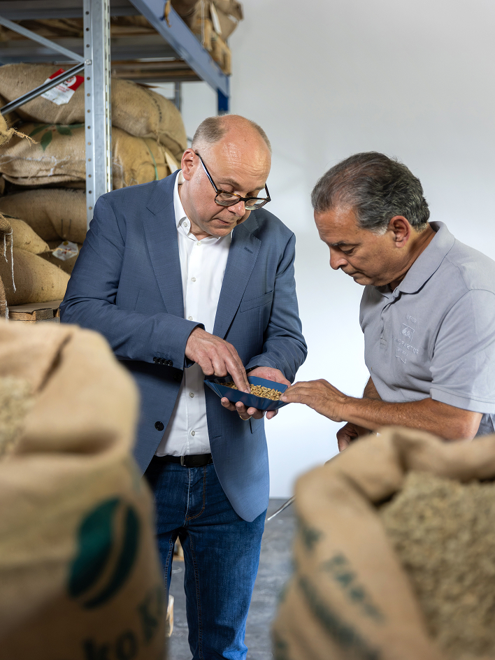 Two men inspect unroasted coffee beans together in a tray among stacked burlap sacks inside a coffee storage warehouse.