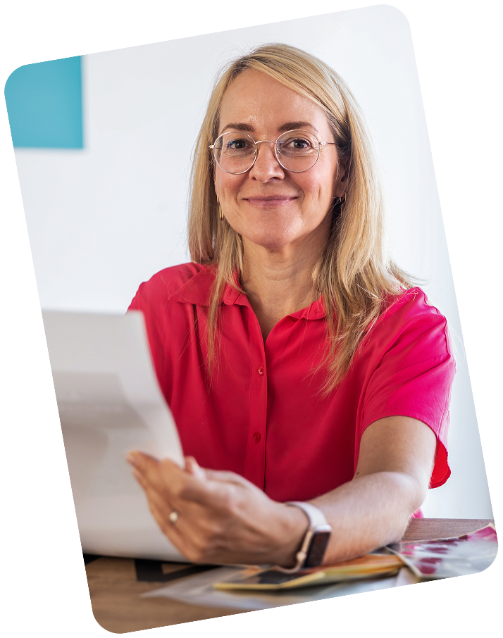 Portrait of the blonde protagonist sitting at a wooden table, holding a design sketch.