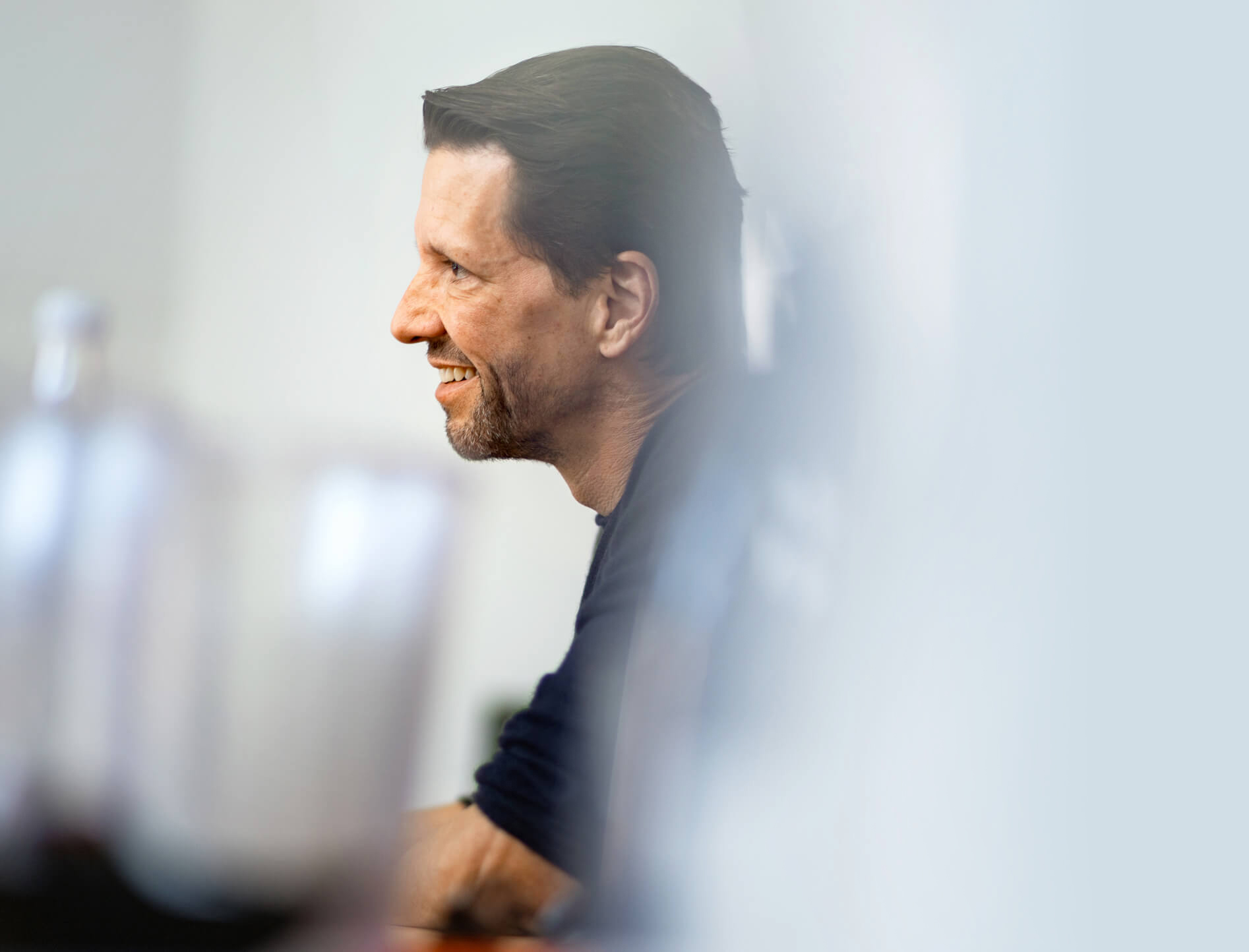 Portrait of Jero Bentz in side profile, framed by blurred foreground elements in a bright room.