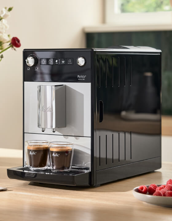 Black automatic coffee machine dispenses two espressos into glasses, with fresh raspberries on the kitchen counter nearby.