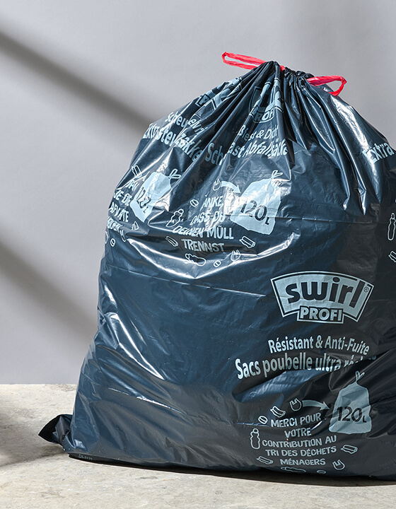 Large black Swirl trash bag with red drawstrings and printed labels rests on a light floor against a white wall.