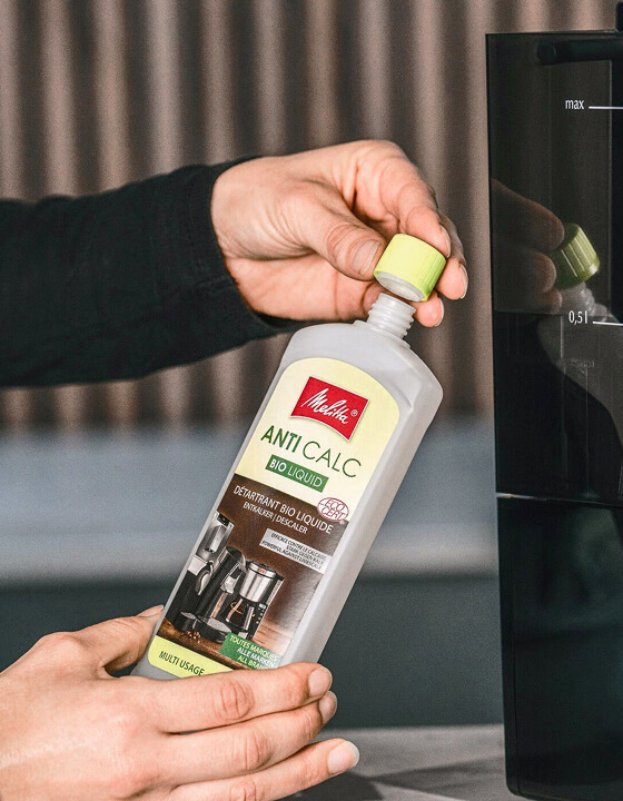 Hands open a bottle of Melitta Bio Liquid descaler beside the water tank of an automatic coffee machine.