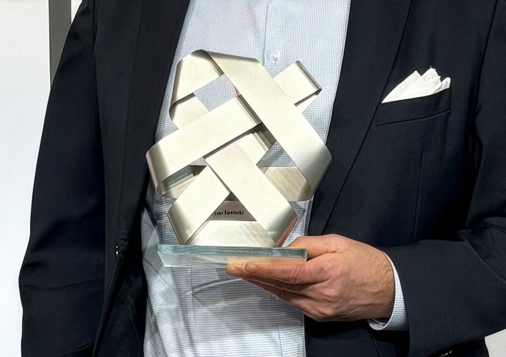 Person in light shirt and jacket holding the silver CSR award sculpture: interwoven metal ribbons on glass base.