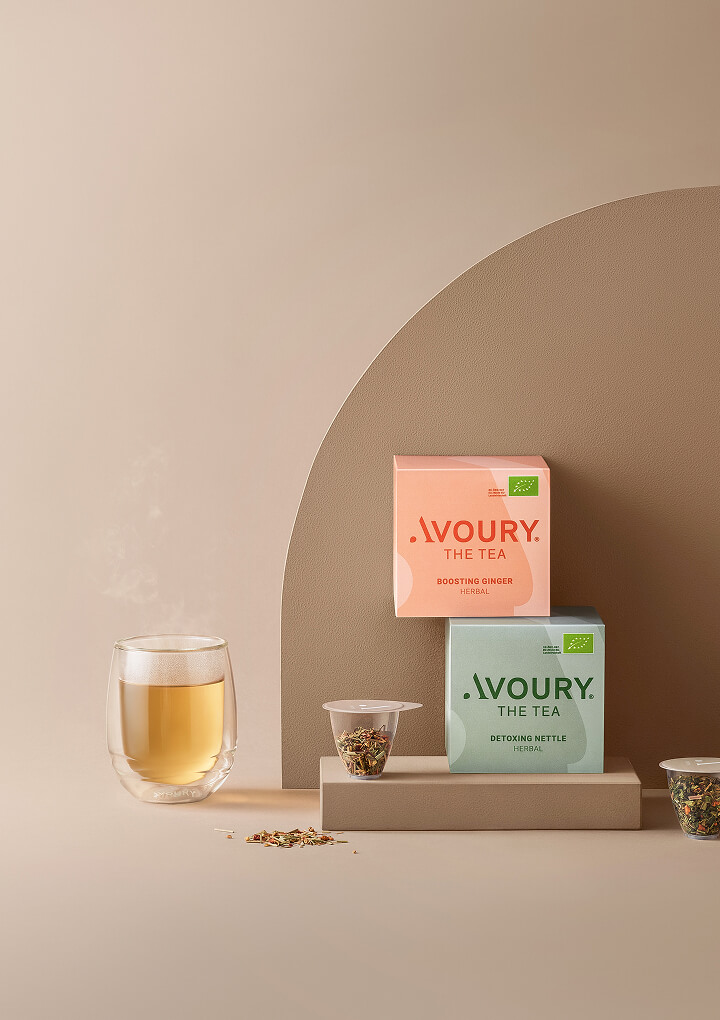 Still life of AVOURY Boosting Ginger and Detoxing Nettle tea packs with capsules and steaming glass cup on beige podium.