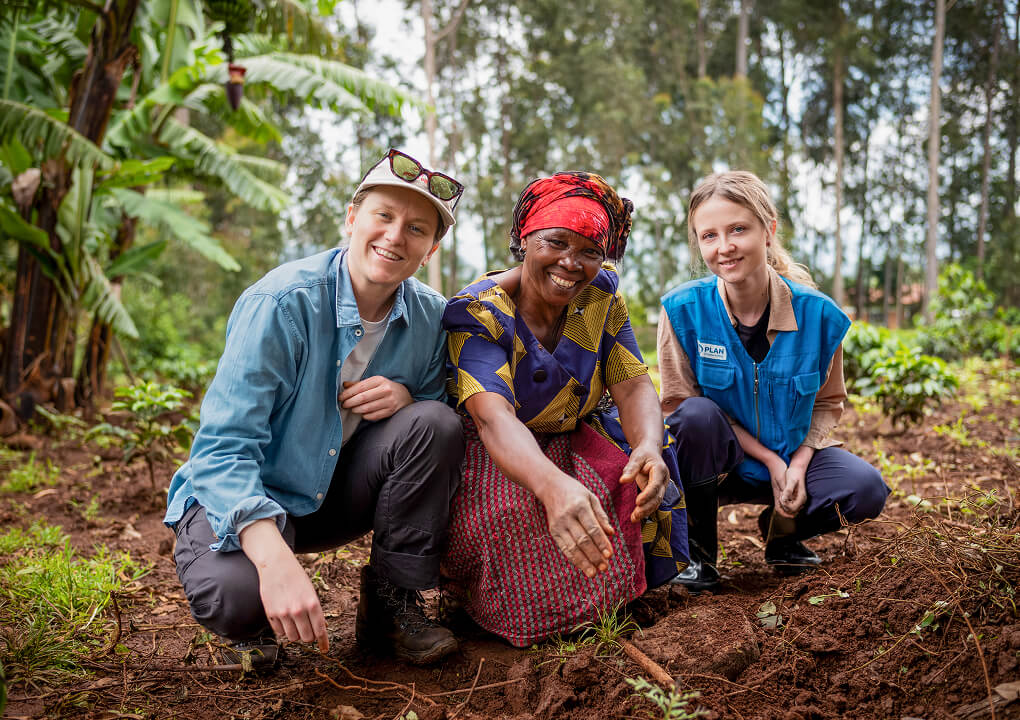 Three women kneel smiling in a coffee plantation field.