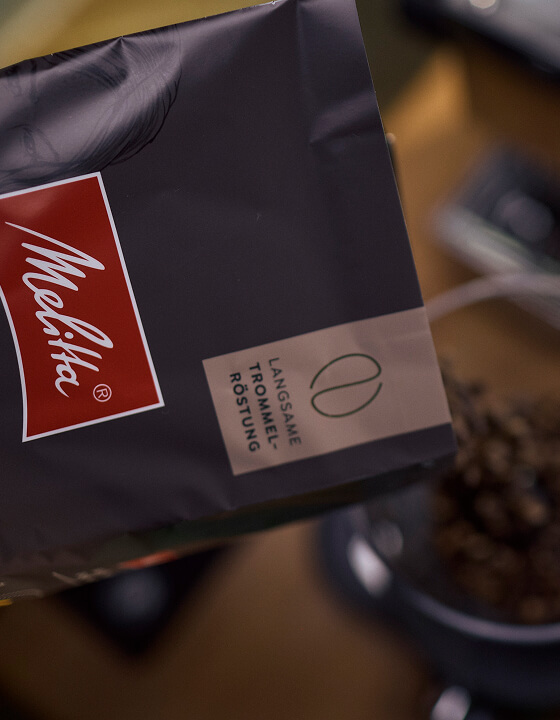 Ge&ouml;ffnete Melitta-Packung mit langsam ger&ouml;steten Kaffeebohnen &uuml;ber einem Beh&auml;lter mit braunen Bohnen.