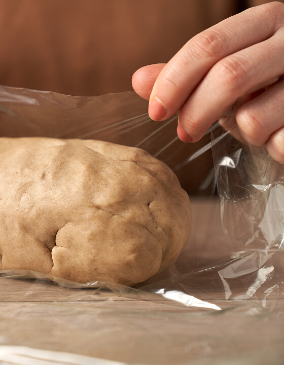 Hand spannt transparente Frischhaltefolie &uuml;ber einer Kugel braunen Teigs auf einer h&ouml;lzernen Arbeitsfl&auml;che.