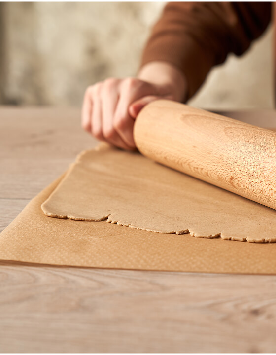 Hand rollt braunen Teig mit einem h&ouml;lzernen Nudelholz auf einer Backmatte &uuml;ber einem hellen Holztisch aus.