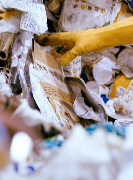 Hand mit gelbem Arbeitshandschuh sortiert bedruckte Kunststoffverpackungen in einem Haufen aus gemischtem Plastikm&uuml;ll.
