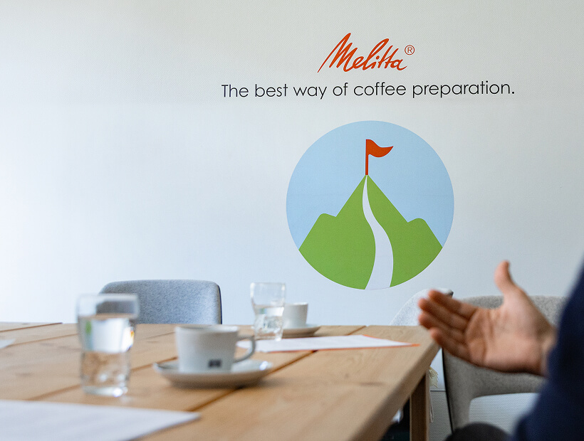 Melitta-Wandbild mit Berggipfel und Text &bdquo;The best way of coffee preparation&ldquo;, davor Besprechungstisch mit Tassen und Gl&auml;ser.