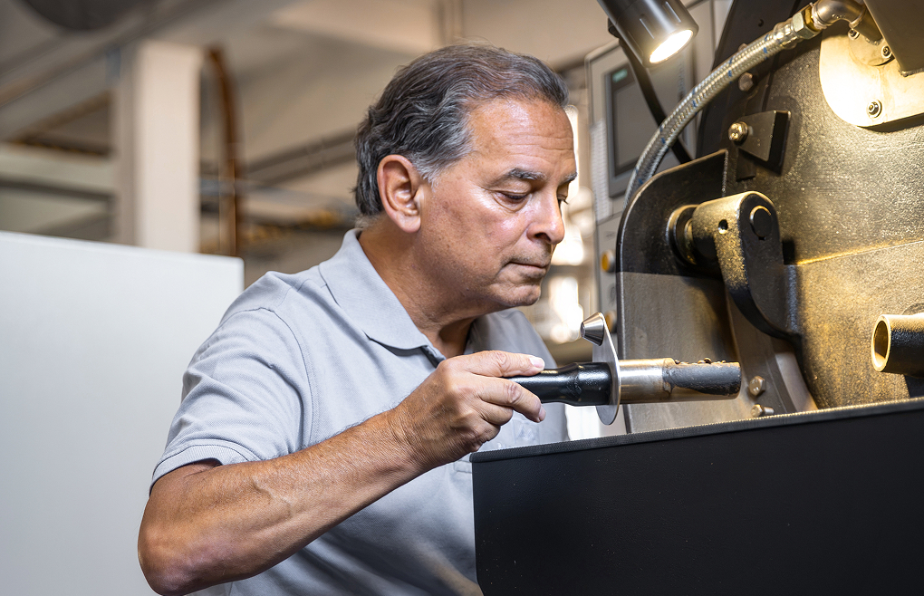 Protagonist entnimmt mit einem Probenehmer ger&ouml;stete Kaffeebohnen aus einer R&ouml;stmaschine zur Qualit&auml;tskontrolle.