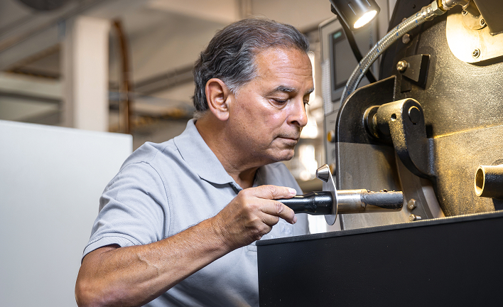 Protagonist entnimmt mit einem Probenehmer ger&ouml;stete Kaffeebohnen aus einer R&ouml;stmaschine zur Qualit&auml;tskontrolle.